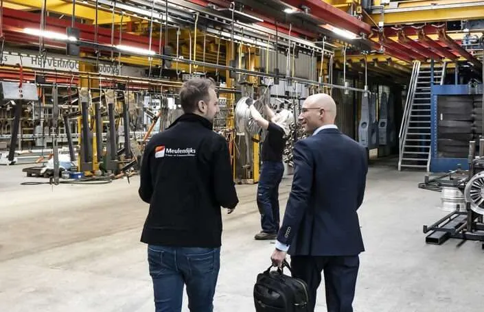 Twee mannen lopen door een productiehal terwijl een medewerker aan het werk is.