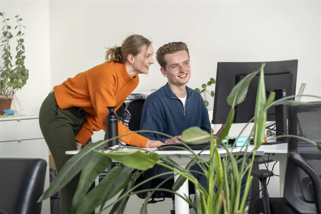Twee collega’s glimlachen terwijl ze samen achter een computer werken op kantoor.