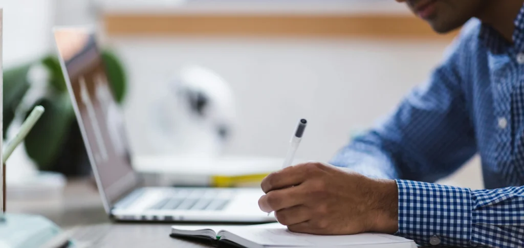 Man maakt aantekeningen in een notitieboek terwijl hij werkt op een laptop.