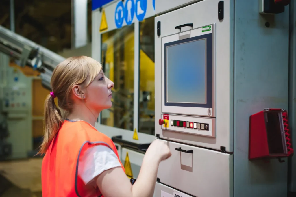Vrouw in veiligheidsvest bedient een industriële machine met een digitaal scherm.